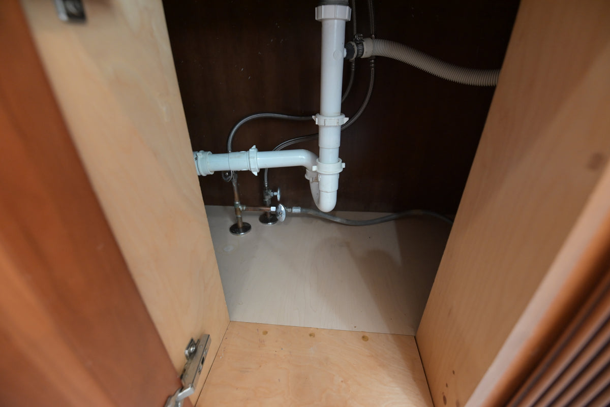 Aurora Circ wooden kitchen cabinet interior with exposed white sink plumbing and open storage base
