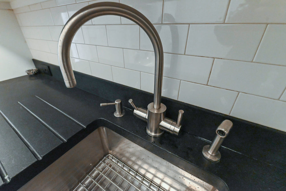 Stainless steel kitchen sink with dual faucet against a white tiled wall.