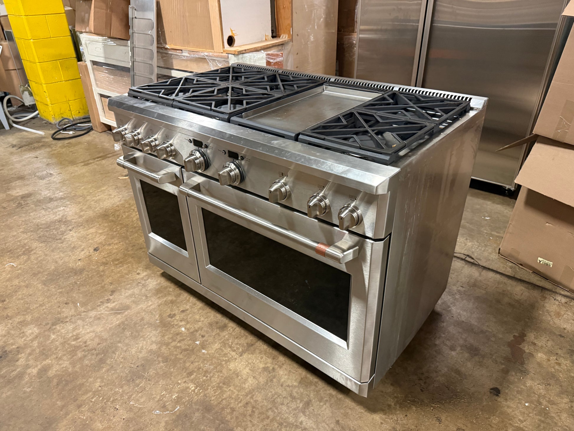 A stainless steel freestanding gas range with a built-in griddle and large front oven doors, photographed in a warehouse or storage area with cardboard boxes and other appliances in the background.