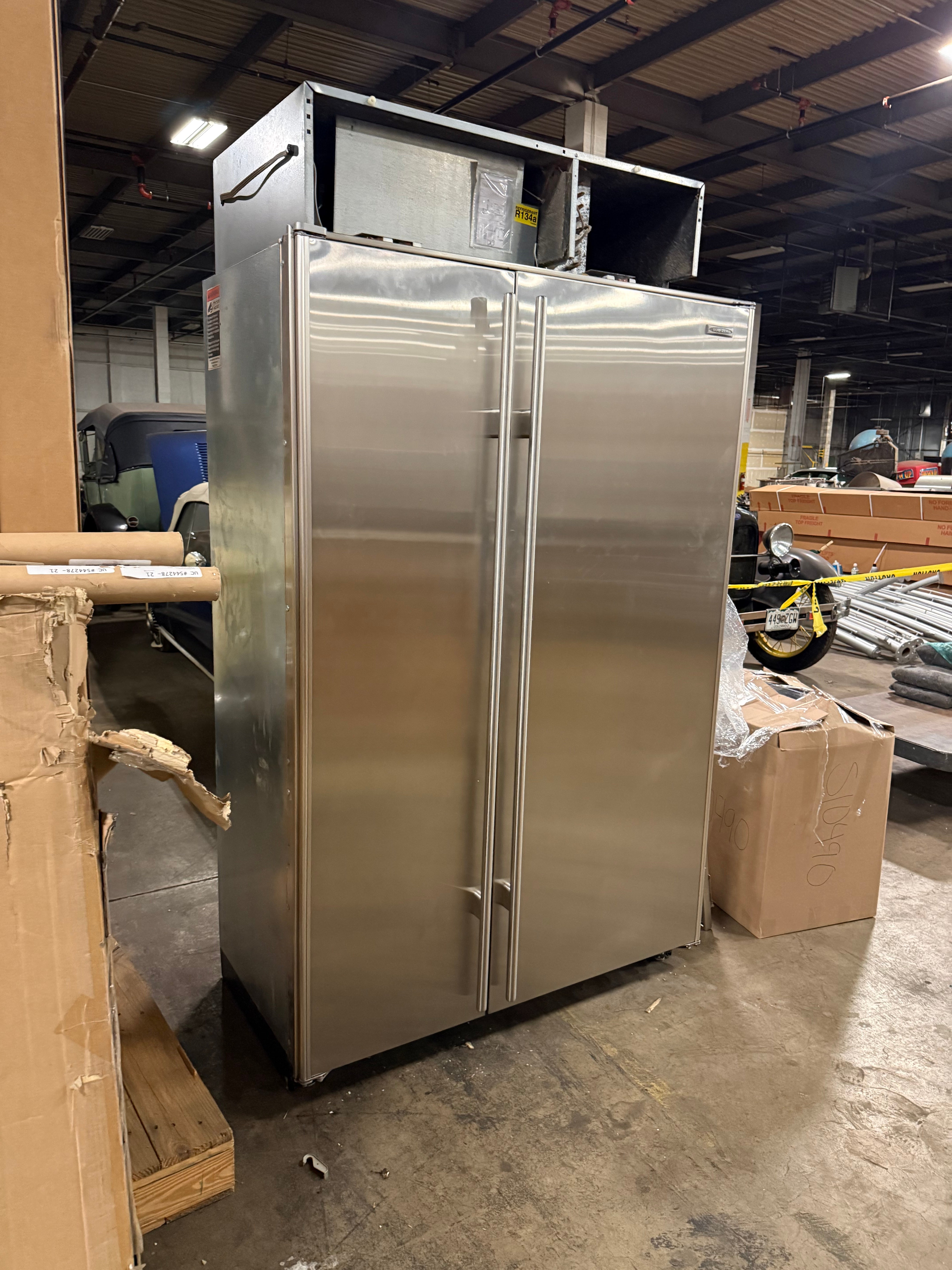 A large stainless steel walk-in refrigerator with two front doors and a top-mounted condenser unit, photographed in a warehouse or showroom setting with boxes and equipment visible around it.