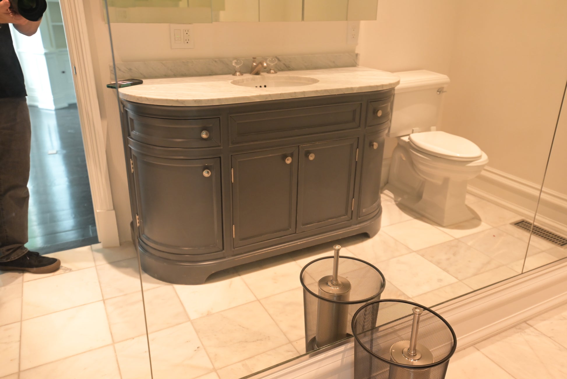 A dark gray bathroom vanity cabinet with a white marble-look countertop and integrated sink, shown in a real bathroom next to a toilet. A person is partially visible on the left edge, and the vanity is reflected in a glass surface in the foreground.