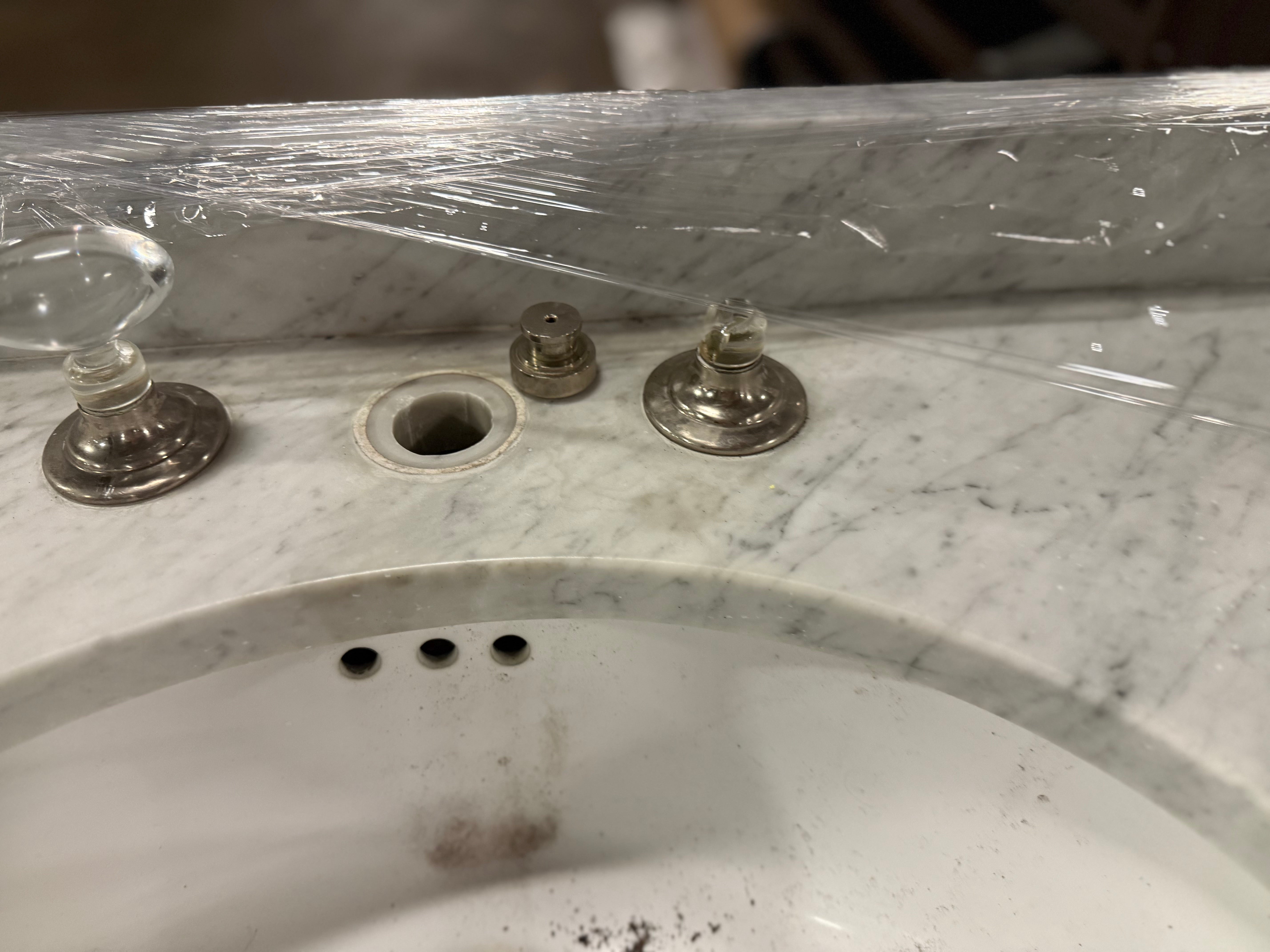 A close-up of a white and gray marble bathroom sink with a missing faucet hole, showing two old metal faucet knobs and a clear glass cap on the left, all covered with protective plastic wrap.